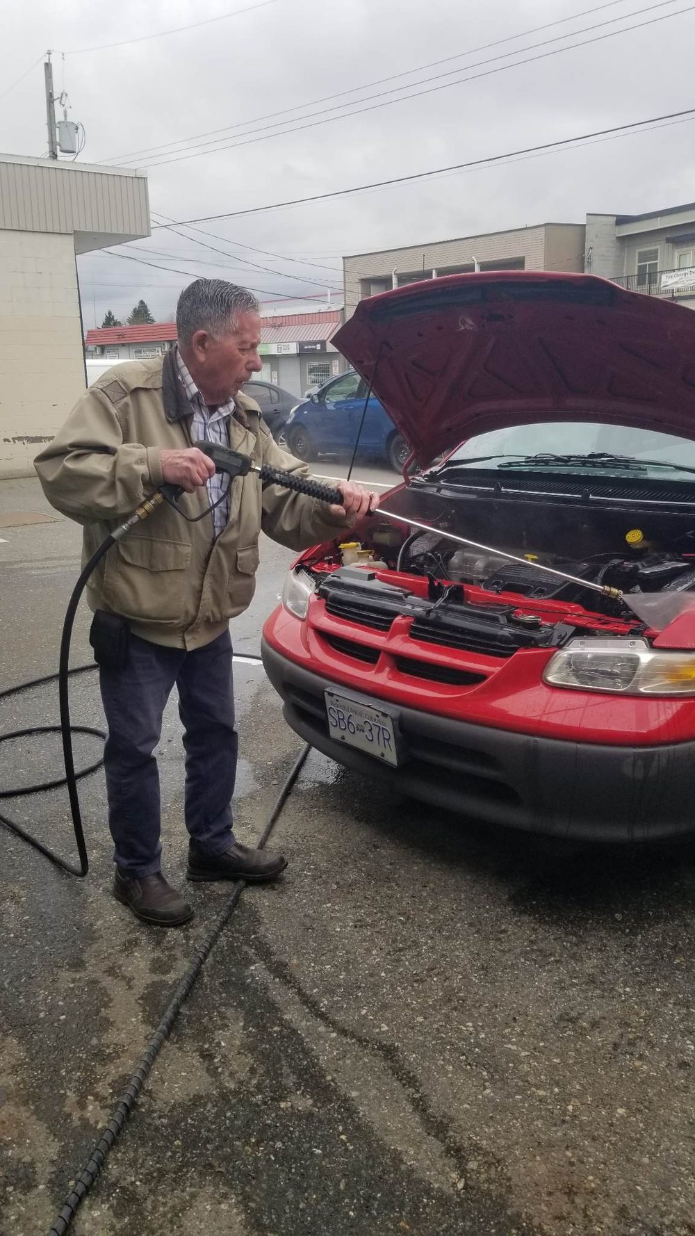 A detailer pressure-washing the engine bay of a red minivan outside the shop in Chilliwack
